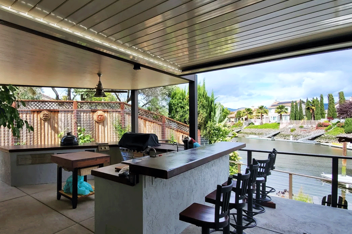 View of a covered outdoor patio with a bar counter, bar stools, and a scenic view of a river, houses, and lush greenery in the background.