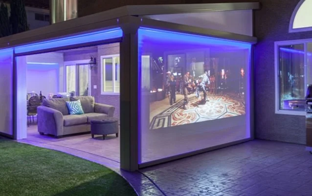 Outdoor patio with a modern pergola featuring a translucent screen showing a movie or TV scene. The area is illuminated with blue LED lights, and there's a sofa with a patterned cushion. Adjacent dining set visible inside the structure.