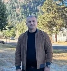 Man standing outdoors in a jacket with a scenic background of trees and hills.