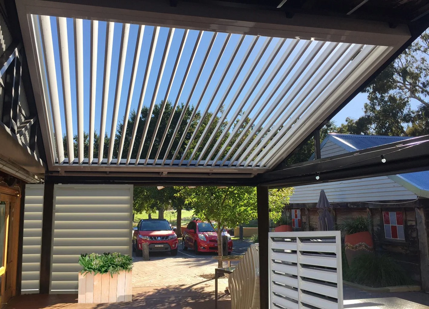 Outdoor patio with adjustable louvered roof and decorative screen, overlooking parked cars and trees.