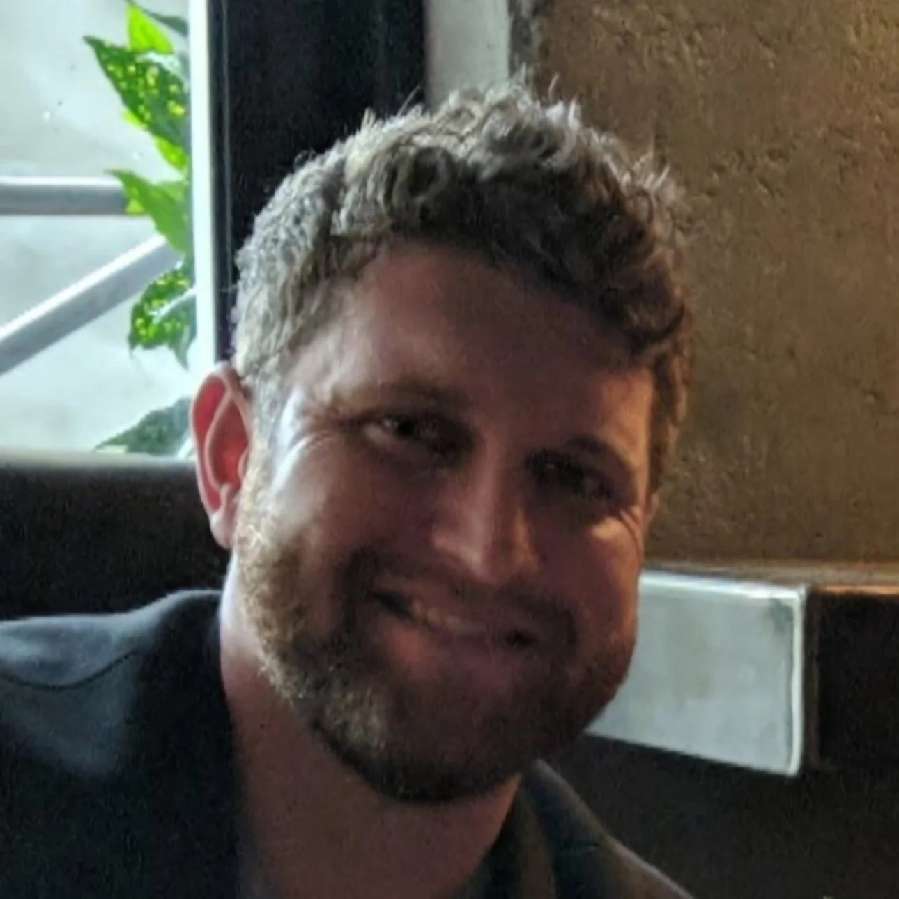 Person smiling indoors with partial view of window and plants