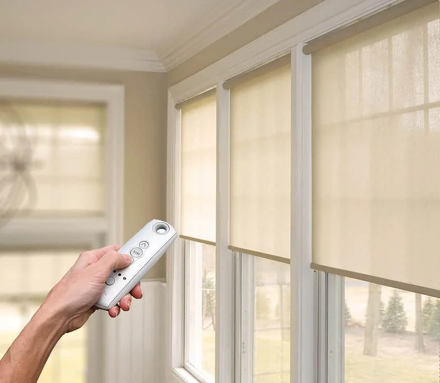 Person using a remote control to operate motorized window blinds in a room with beige walls.