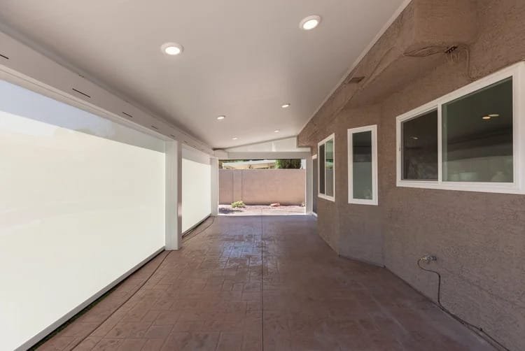 Covered patio with stamped concrete floor, white ceiling with recessed lighting, and exterior windows; view of a wall with a garden bed outside.