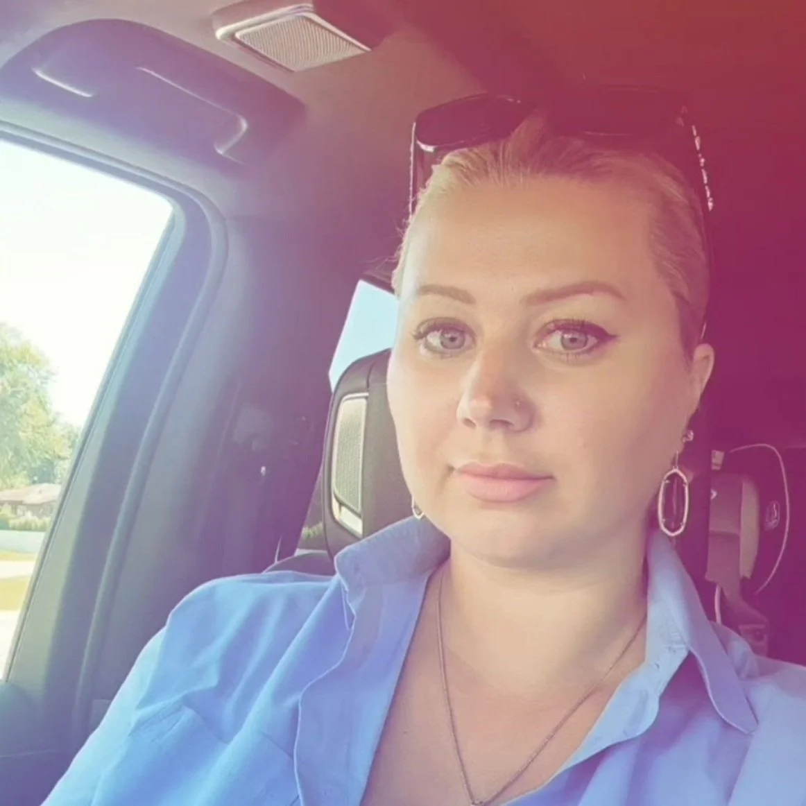 Woman with short hair wearing a blue shirt, sitting in a car, looking at the camera.