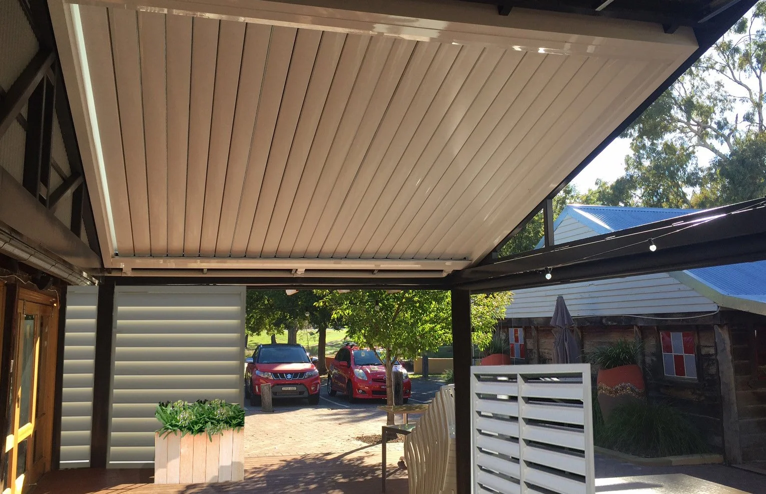 Outdoor patio with louvered roof system, two red cars in background, wooden deck with plants, and a view of trees and buildings.