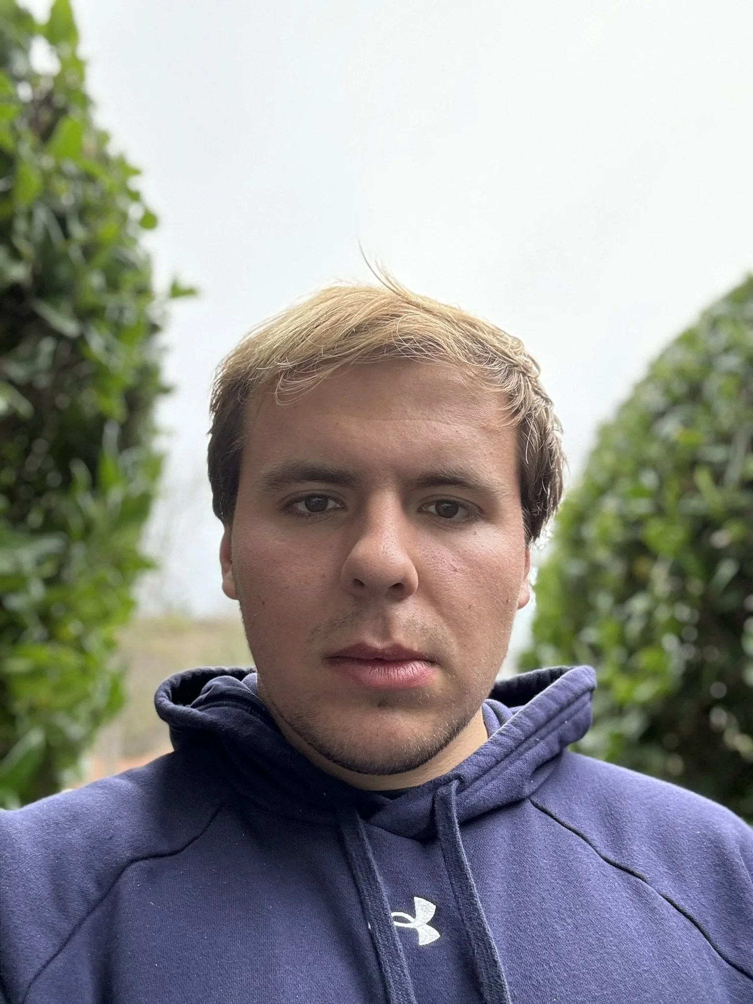 Person wearing a blue hoodie standing outdoors with green foliage in the background.