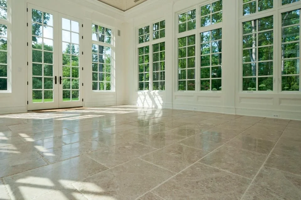 Spacious sunroom with large white-framed windows and French doors, beige tiled floor, and view of green trees.
