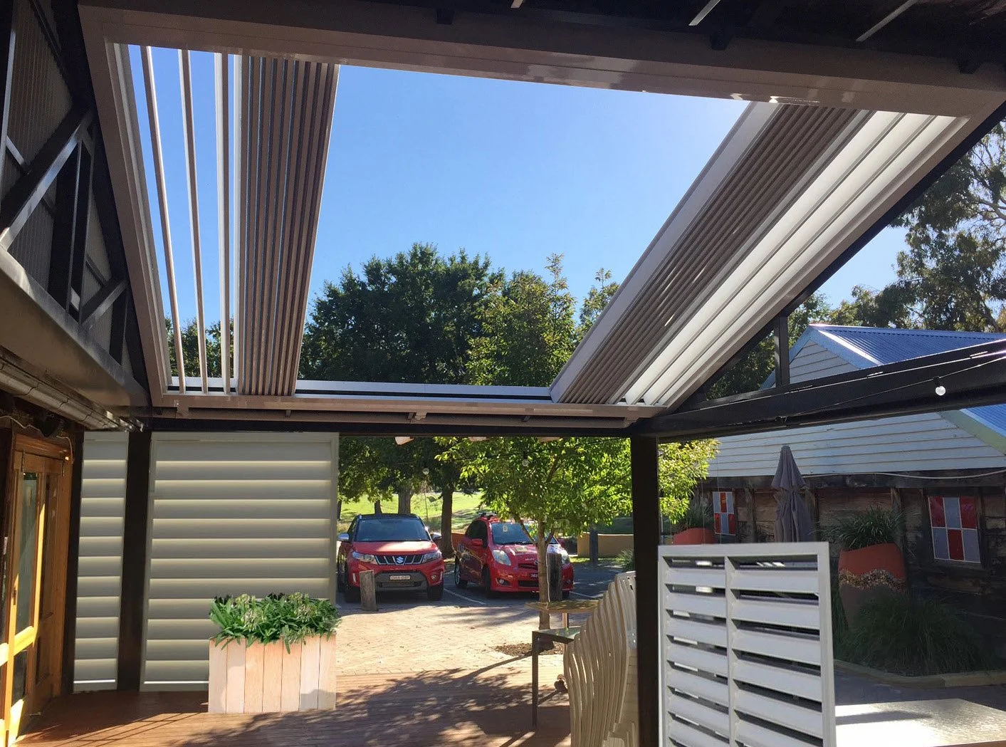Outdoor patio with pergola and louvered roof, cars parked nearby, and trees in the background.