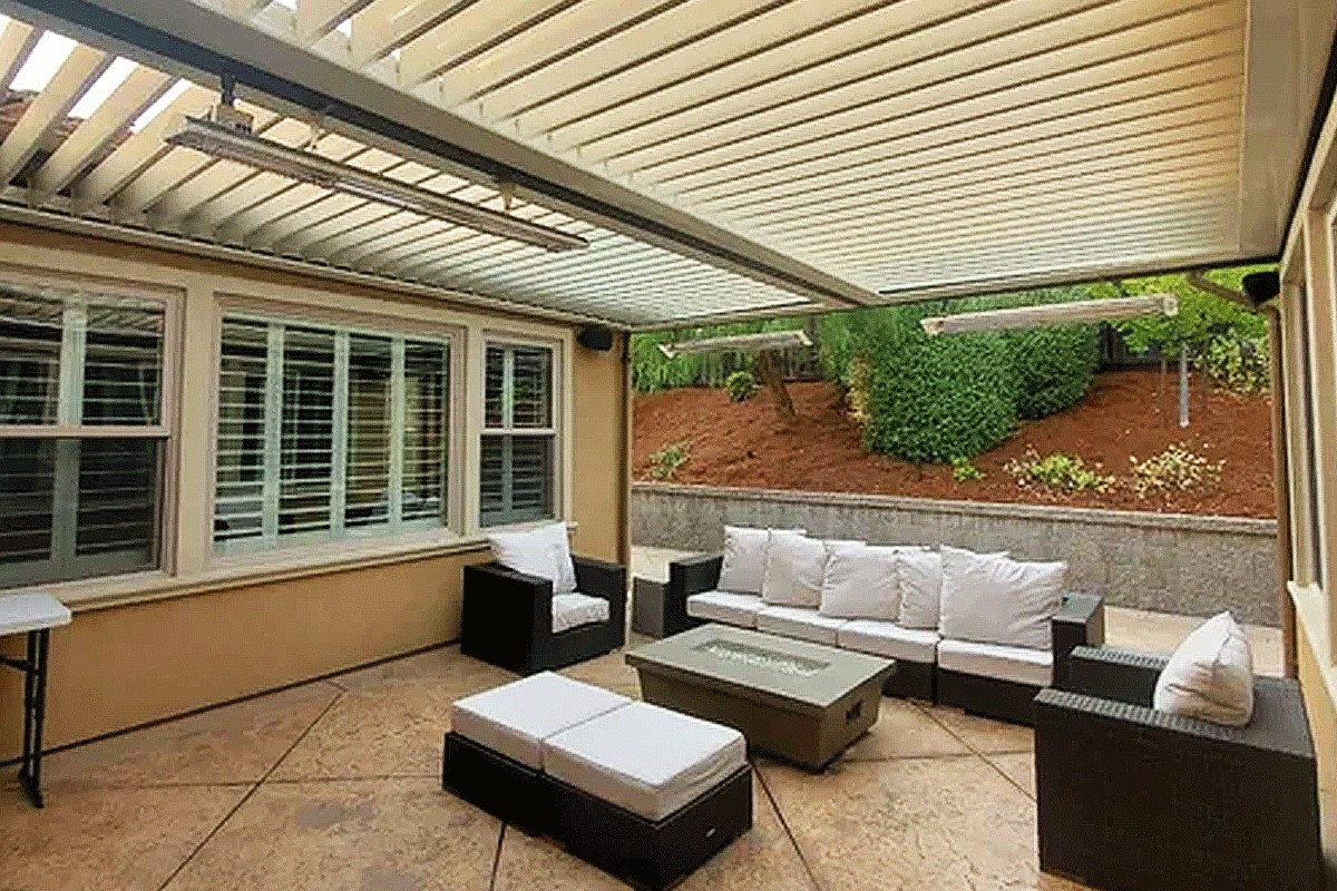 Covered outdoor patio with ceiling fans, a large granite kitchen island with a sink, dining table with chairs, and a seating area with couches and potted plants, surrounded by a wooden fence with garden beds and colorful flowers.