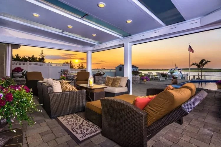 Outdoor patio with wicker furniture, a table with a lantern, vibrant cushions, and flower pots against a waterfront view at sunset.