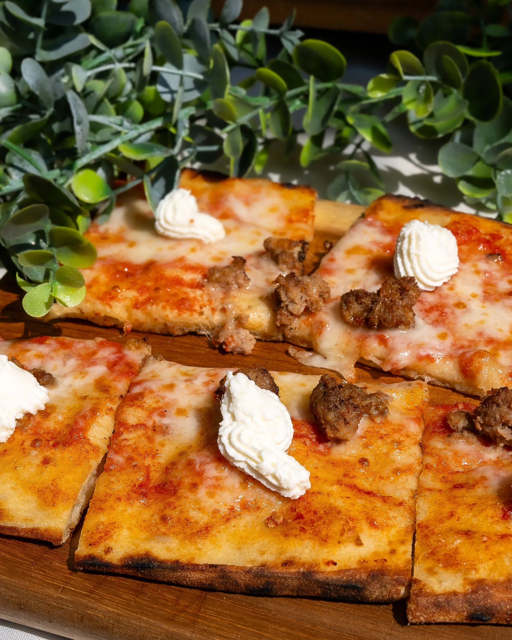 Slices of cheese pizza with dollops of white cheese and crumbles of sausage on a wooden platter, surrounded by green foliage.