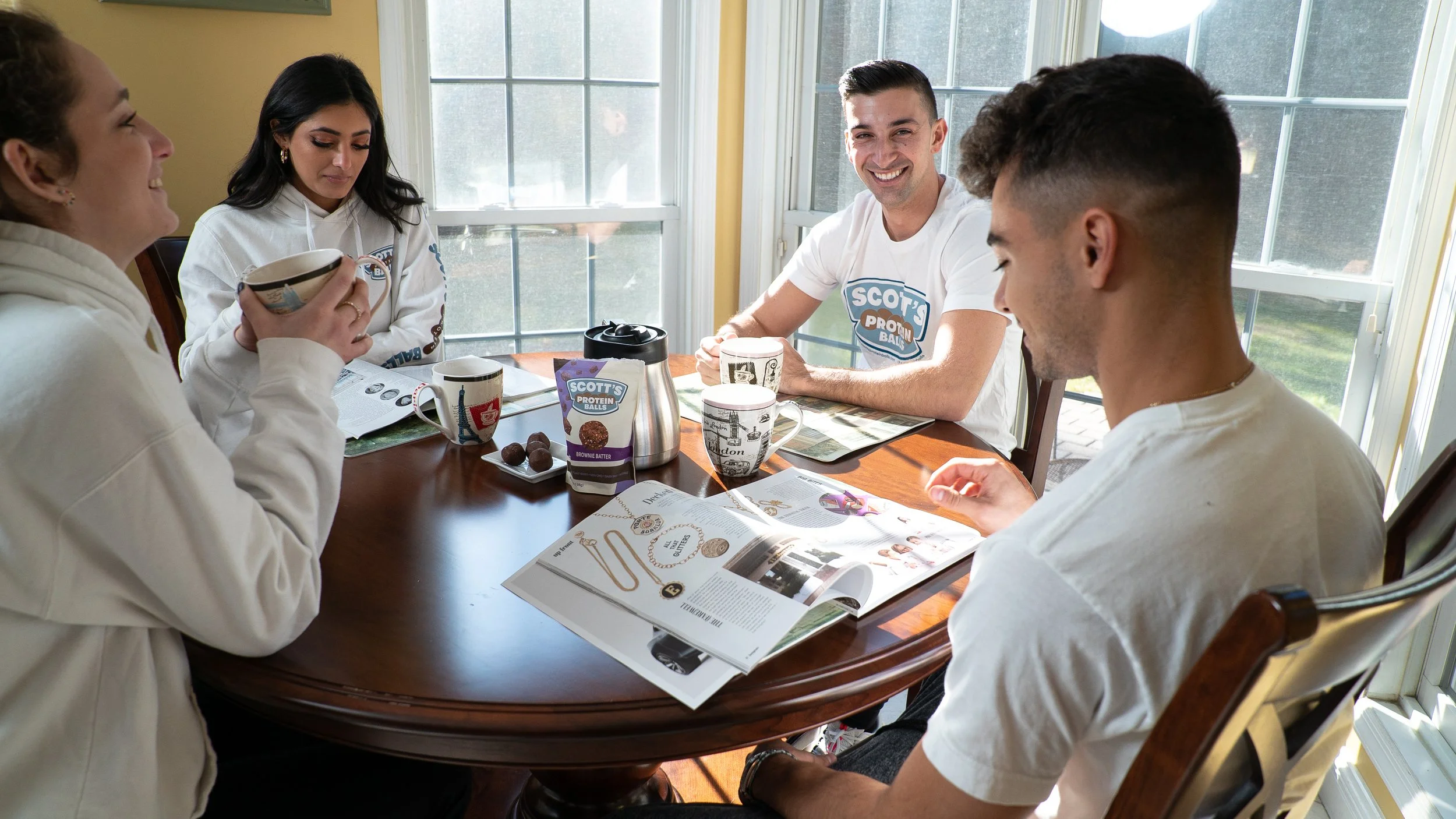 Five young adults sitting around a wooden dining table, smiling and chatting during breakfast time, with magazines and cups of coffee on the table.