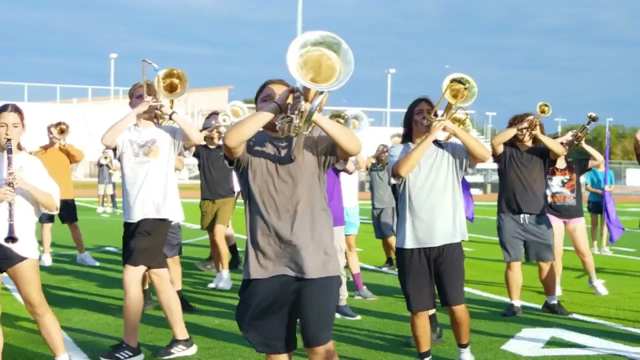 A group of young people playing brass and woodwind instruments in a marching band practice on a football field.