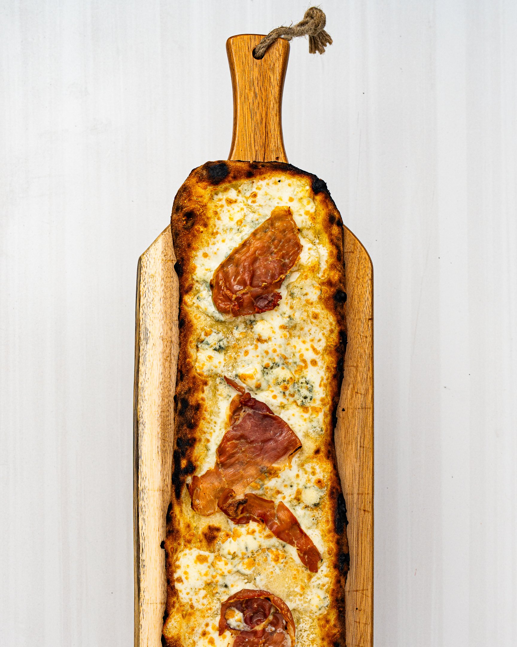A rectangular pizza with cheese and slices of prosciutto, placed on a wooden serving board with a handle and string, on a white surface.