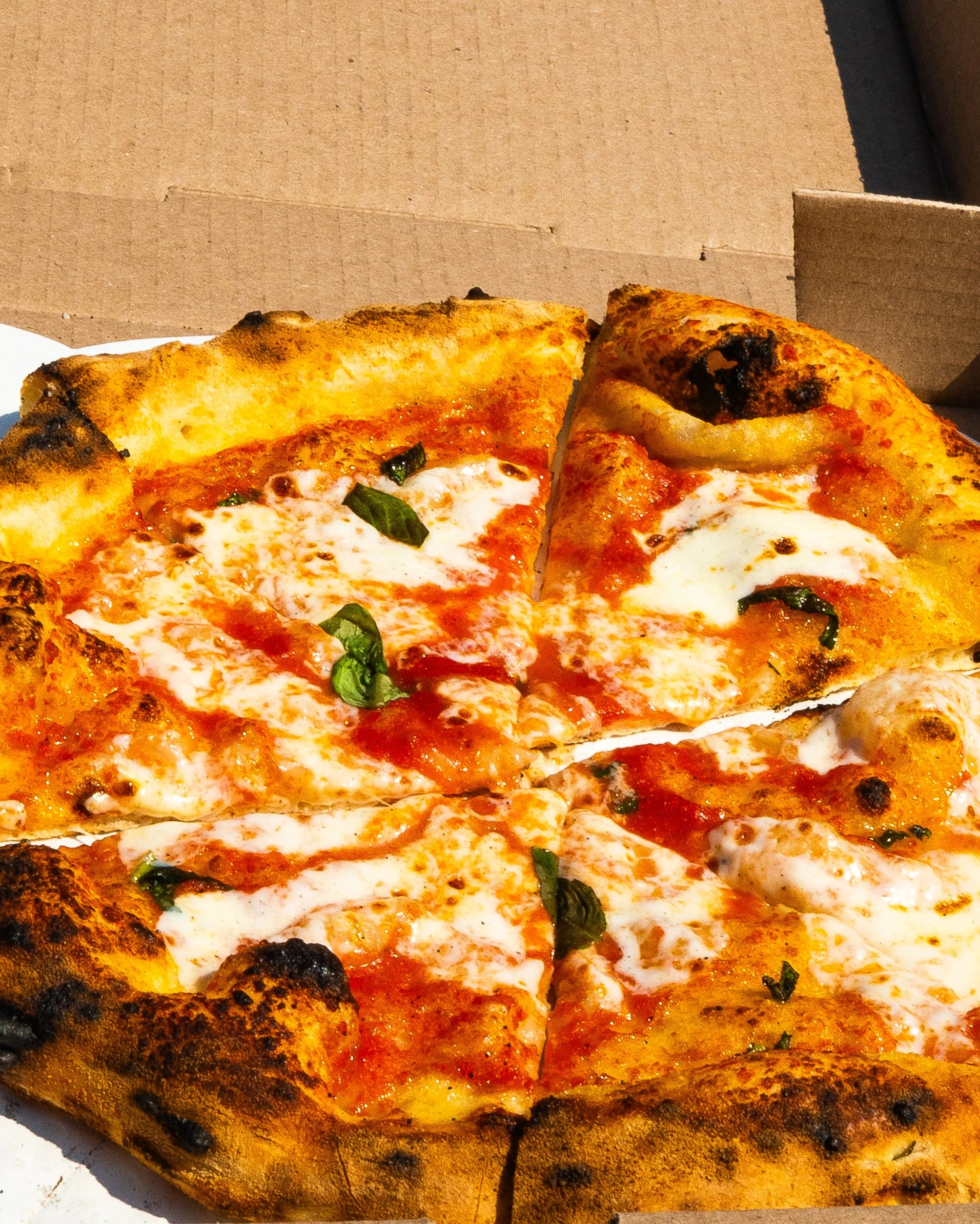 Slice of Margherita pizza with cheese, tomato sauce, basil, and a slightly charred crust, in a pizza box.