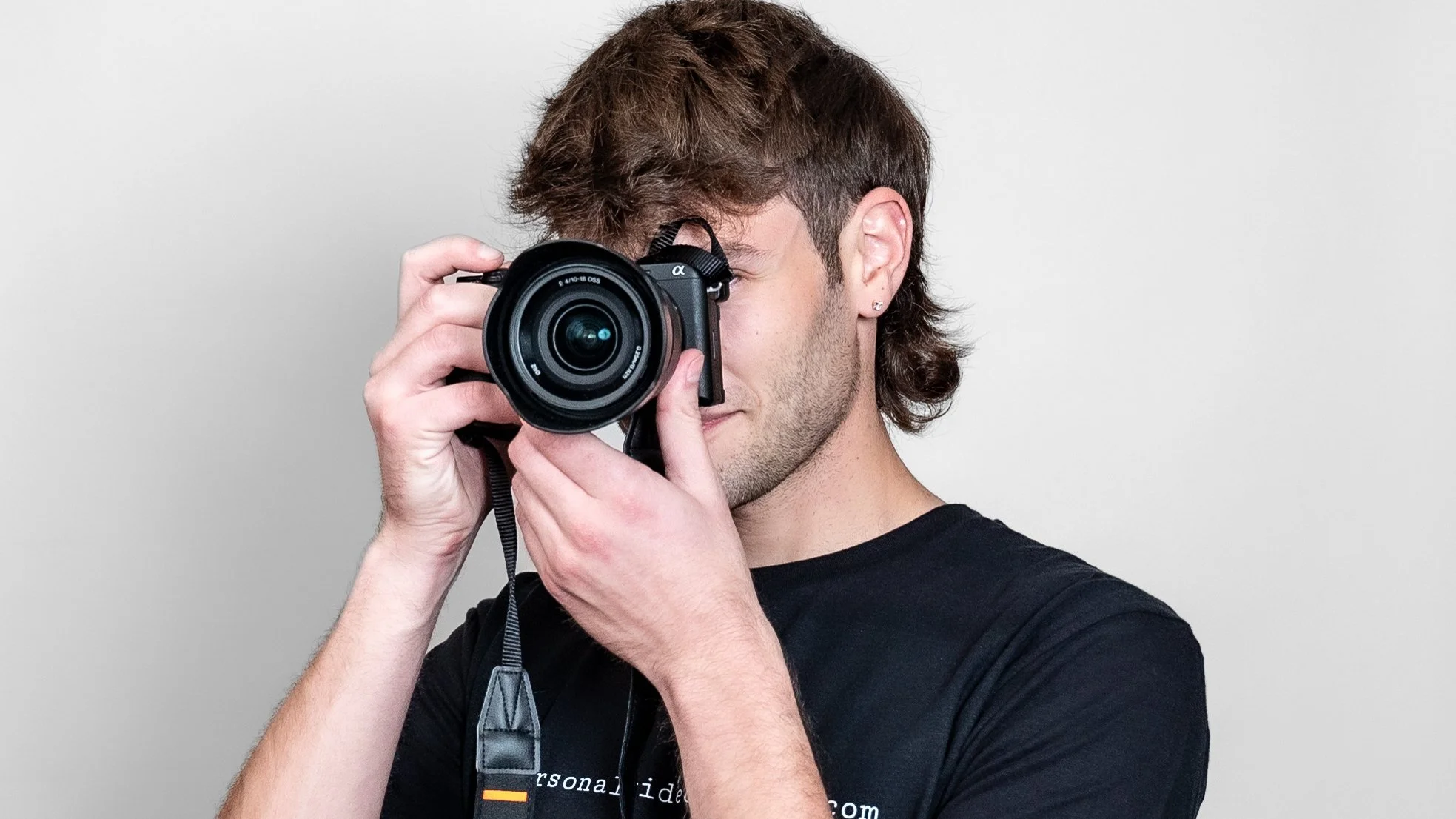 A young man with brown hair and earrings is holding a camera up to his eye and taking a picture against a plain gray background.