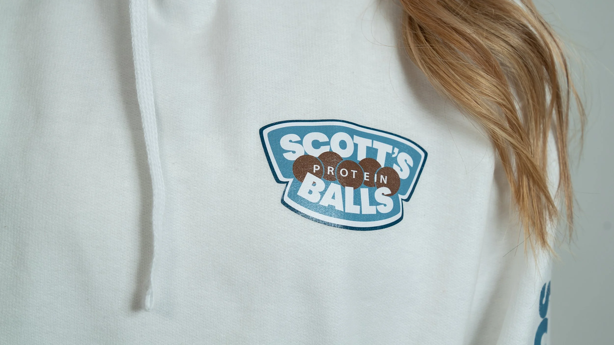 White hoodie with a logo that reads 'Scott's Protein Balls' and features four chocolate protein balls. A person's red hair is partly visible on the right side.