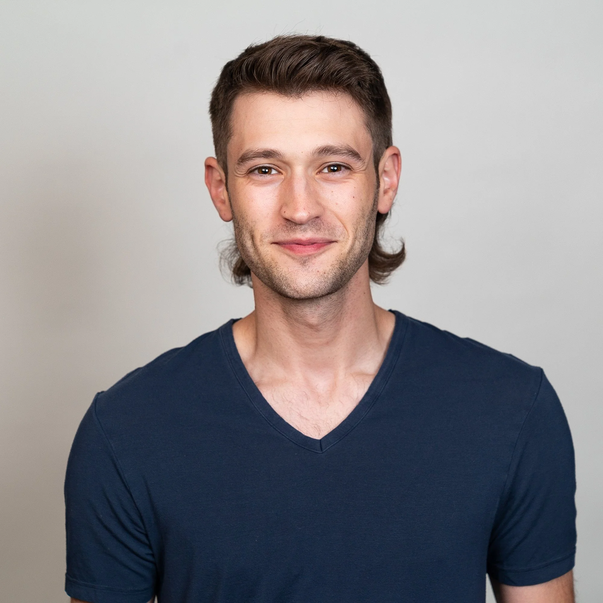 Portrait of a young man with brown hair, smiling, wearing a navy blue V-neck T-shirt, against a plain light gray background.