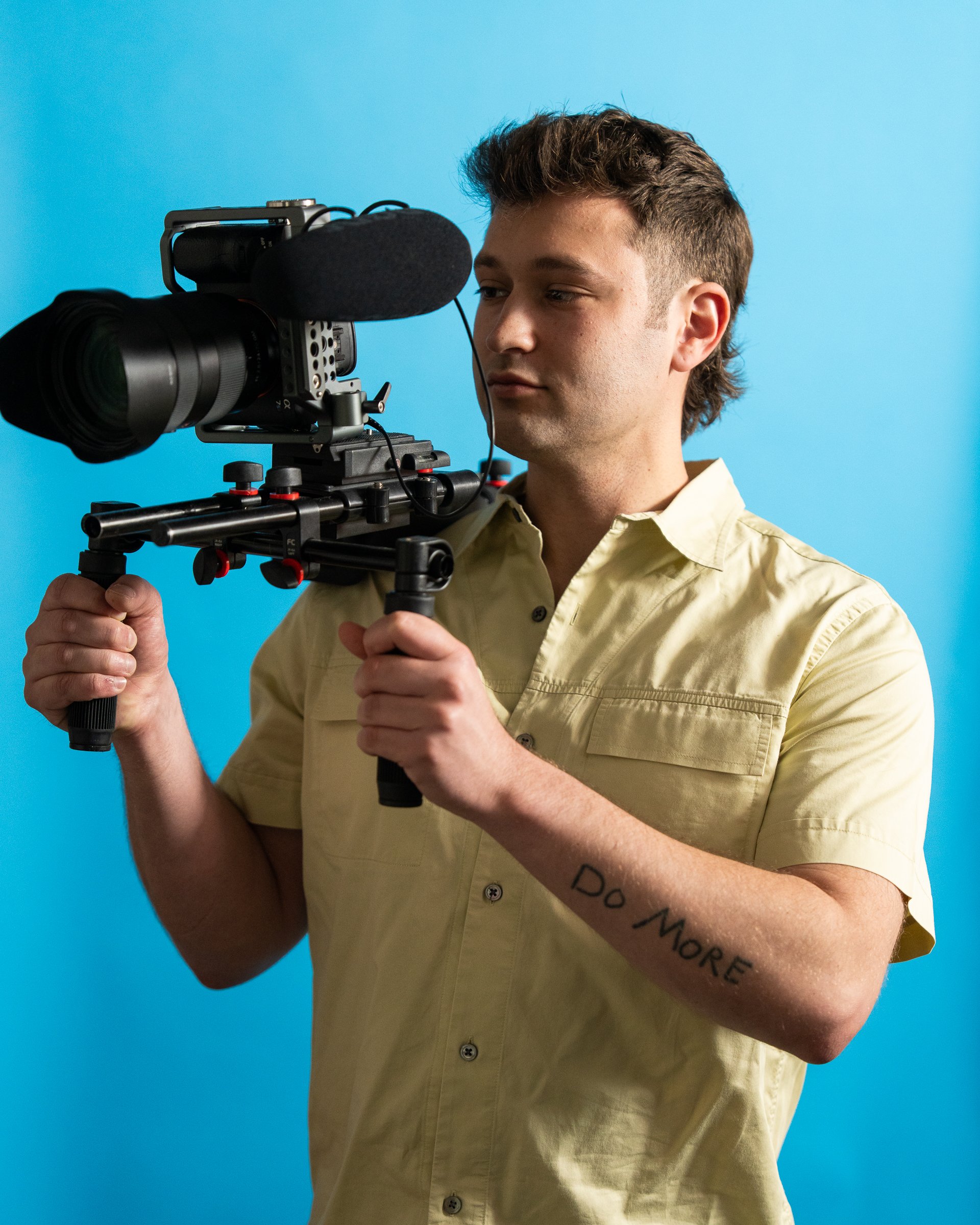 A man in a beige shirt holding a professional video camera against a blue background.