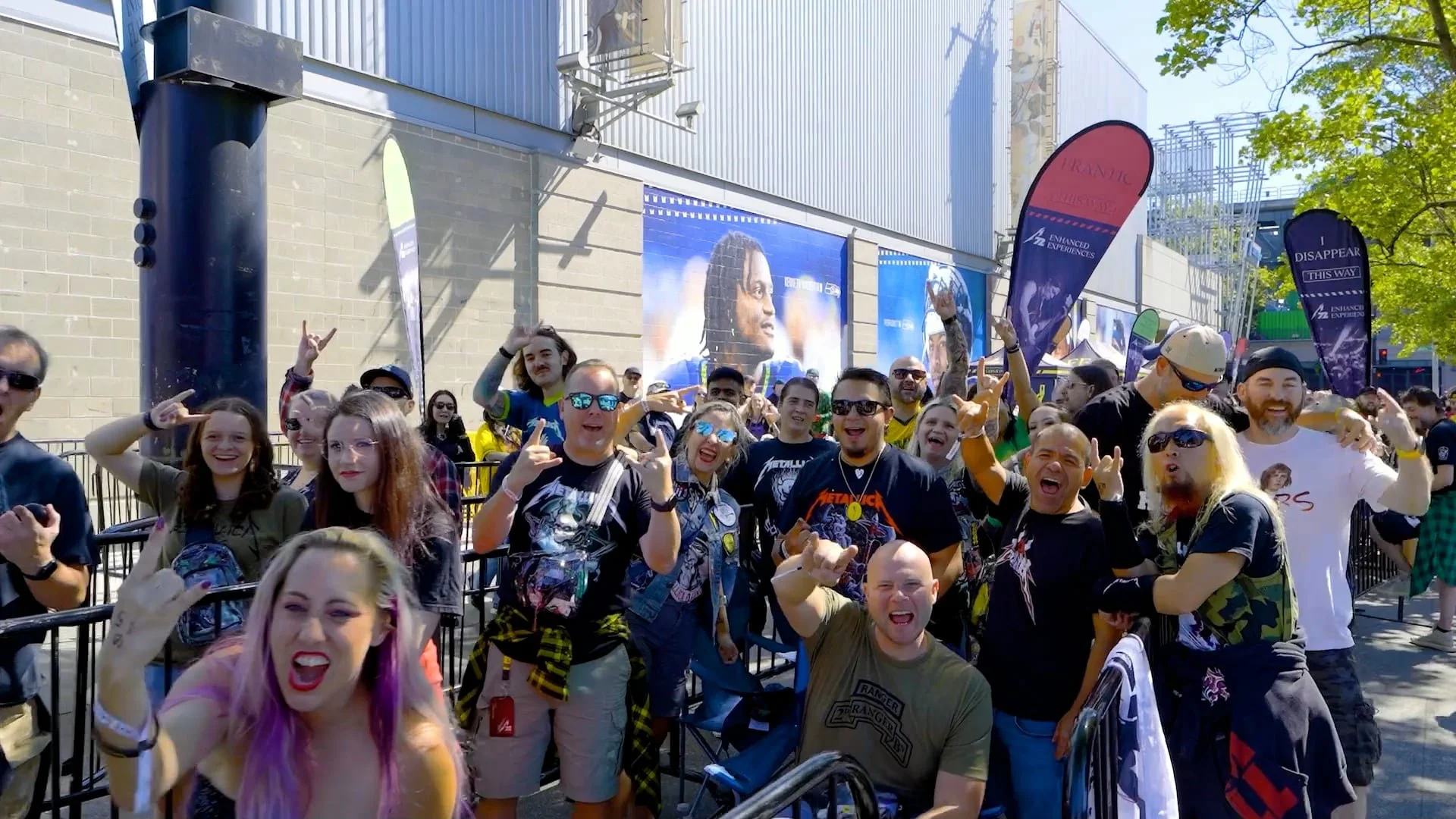A group of people at an outdoor event, many making rock hand gestures and smiling. Some are wearing metal band t-shirts, sunglasses, and casual clothing. There are banners and posters in the background, including one with a football player and some t