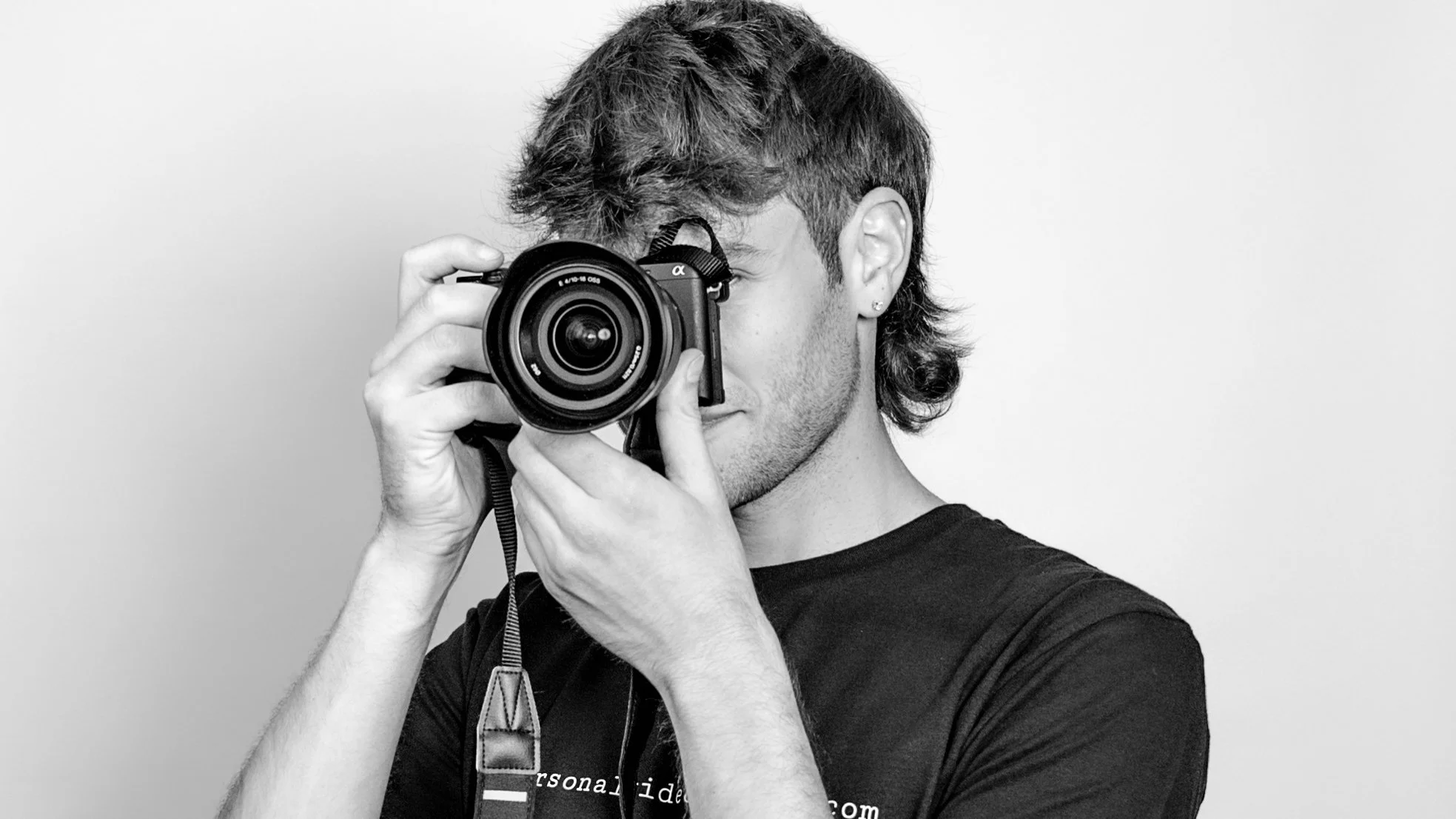 A young man with brown hair and earrings holding a camera up to his face, taking a photo, wearing a black T-shirt, in a black and white photo.