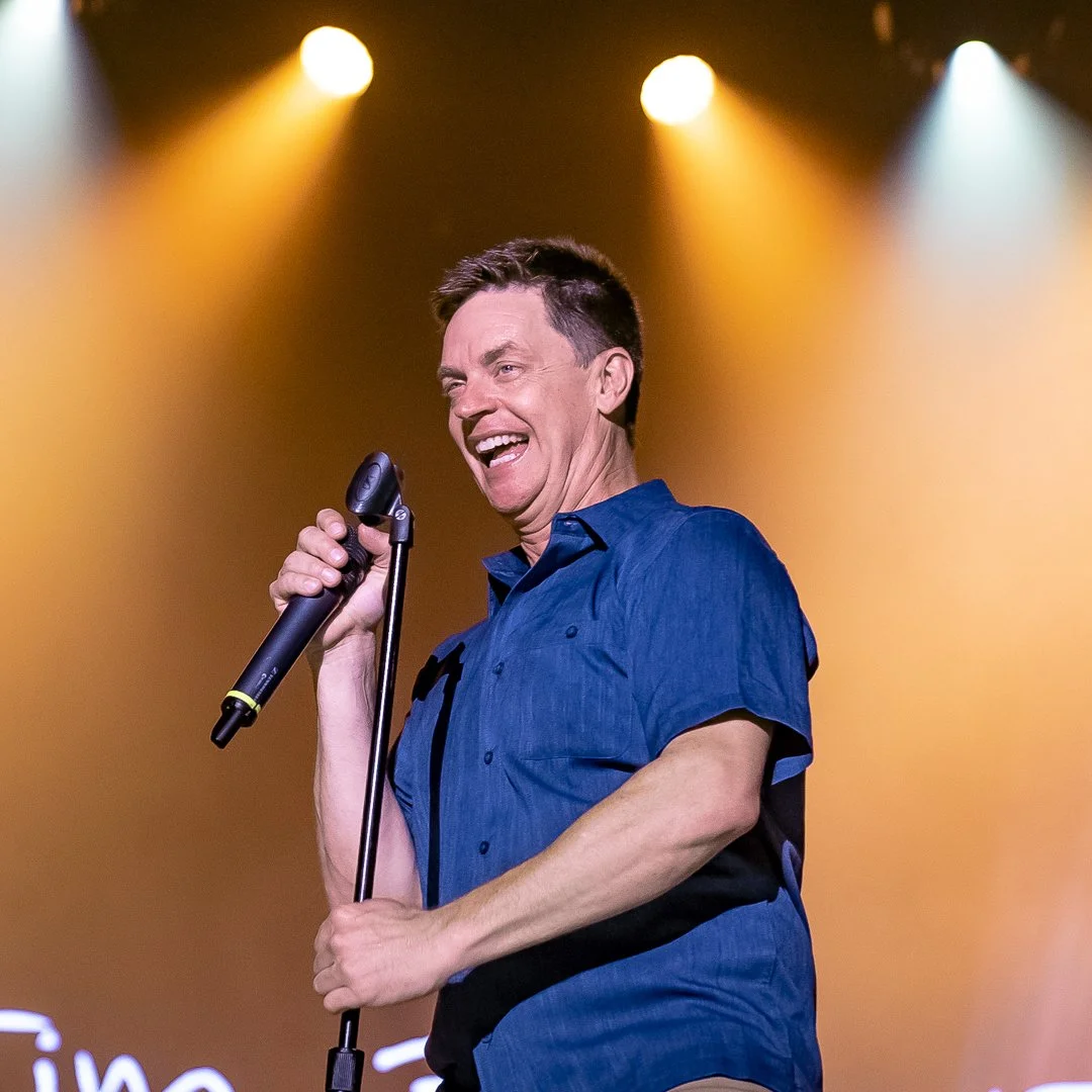A man singing into a microphone on stage with colorful lighting in the background.