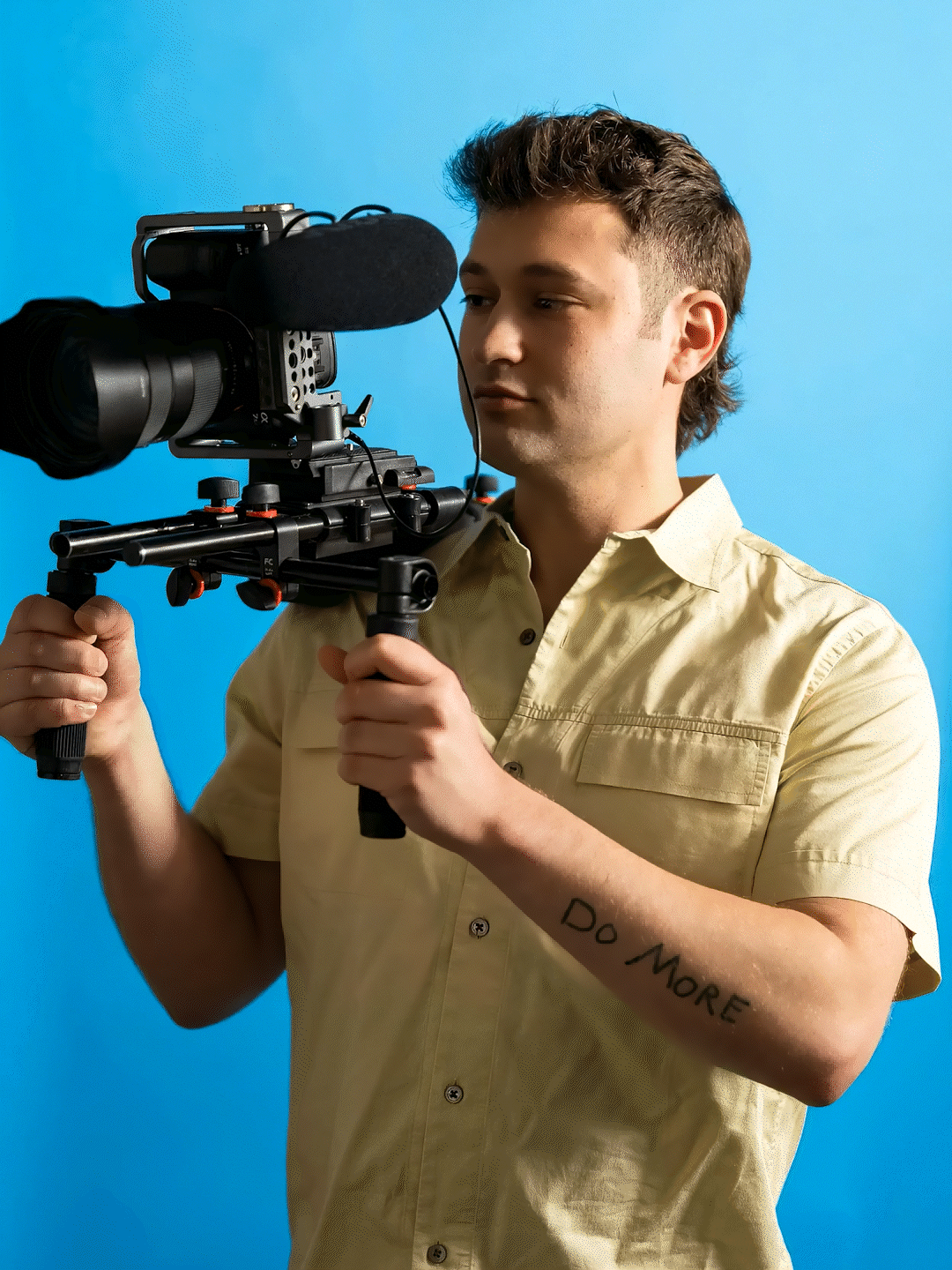 A young man with a tattoo that says 'Do More' on his right arm holding a professional camera with a microphone against a blue background.