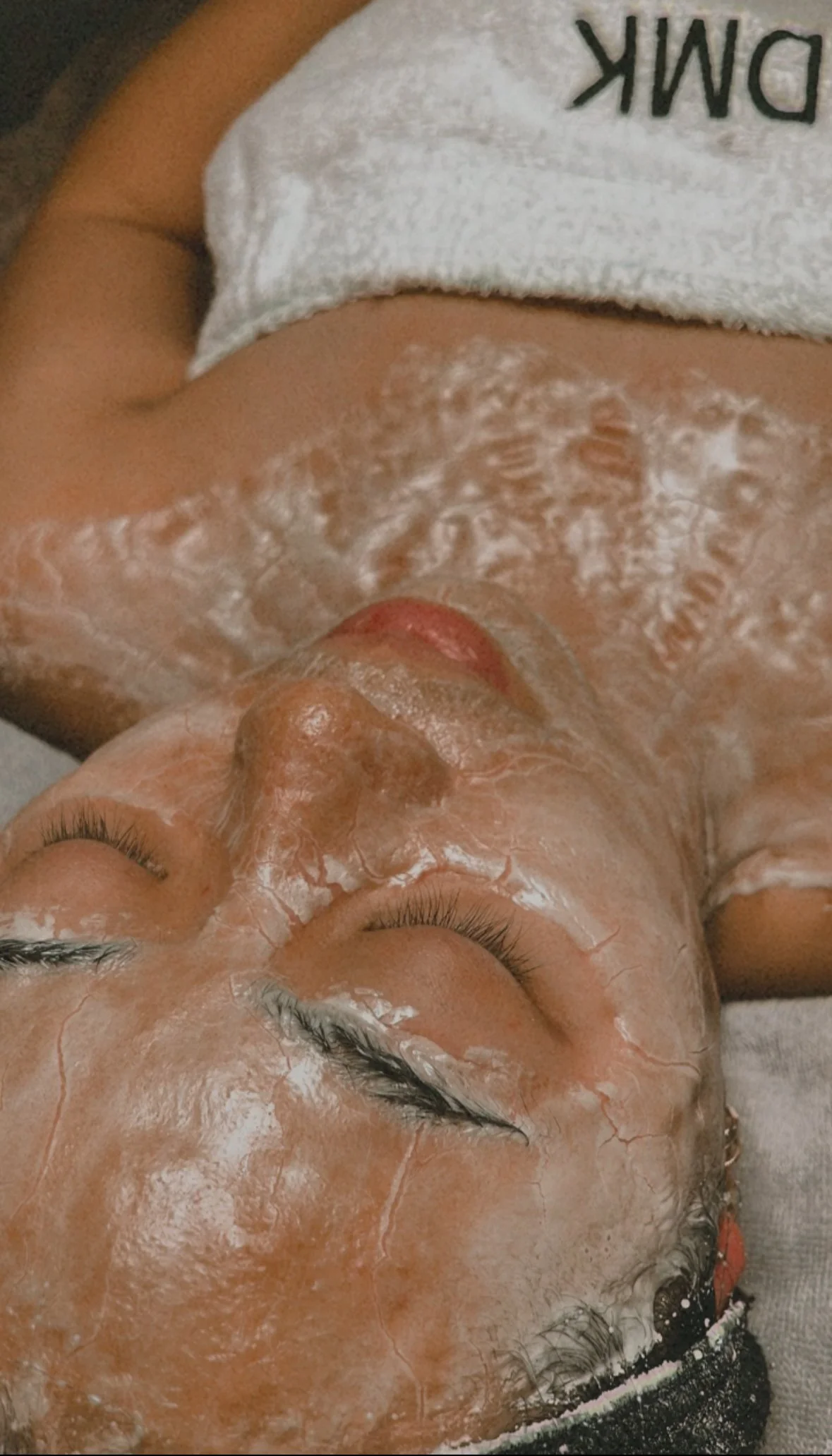 Person receiving a facial treatment with DMK enzyme mask applied, eyes closed, towel wrap around her body.