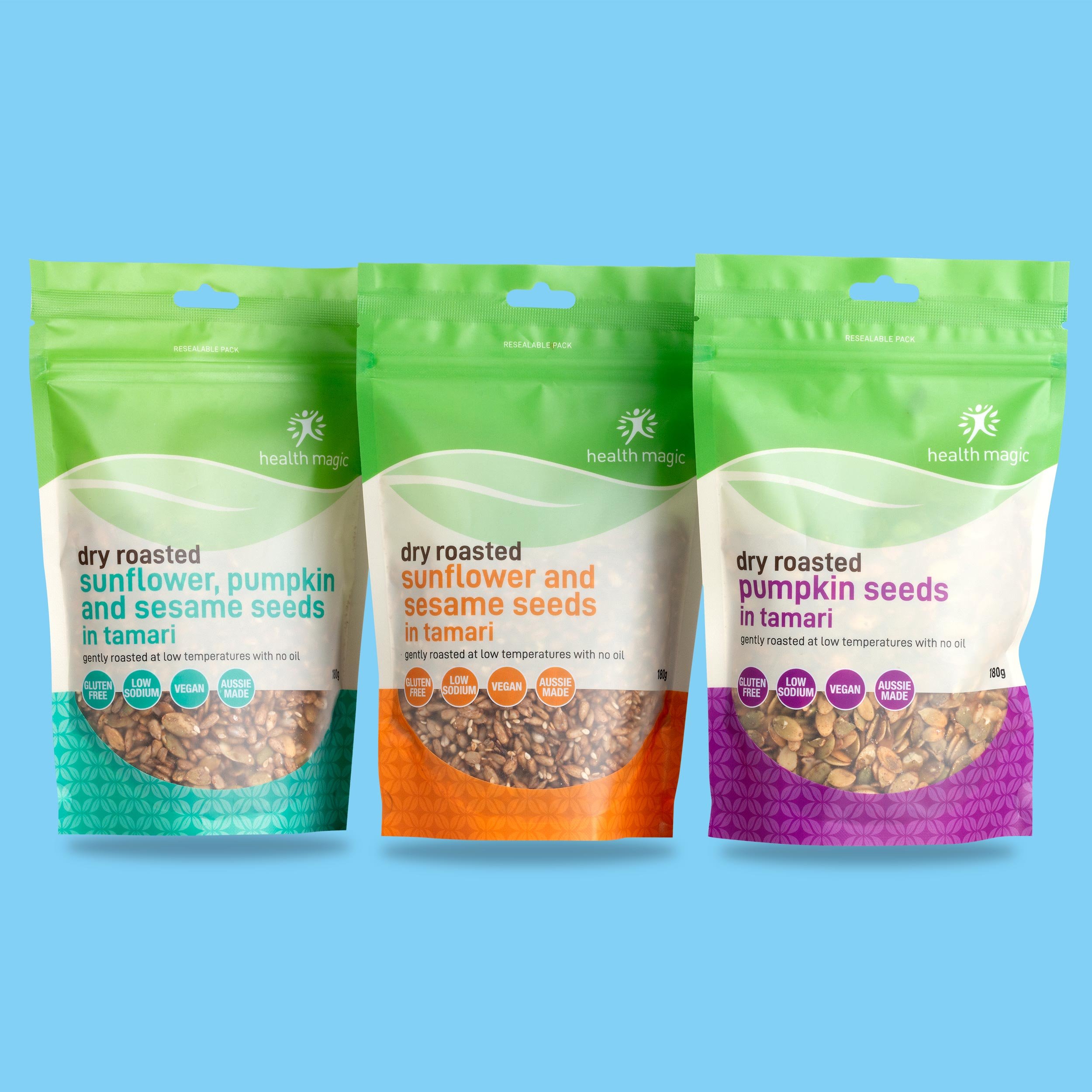 Three resealable bags of dry roasted sunflower, pumpkin, and sesame seeds in tamari flavor, with one bag of each flavor arranged in front of a blue background.