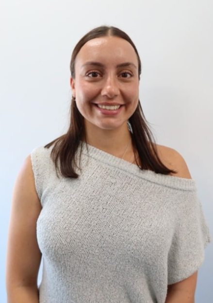 Close-up portrait of a smiling woman with brown hair pulled back, wearing a white striped top, earrings, and a delicate necklace, standing against a plain light-colored background.
