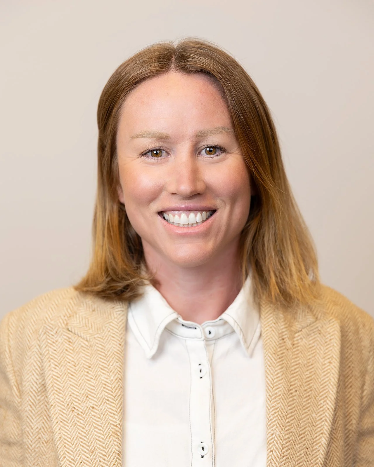A woman with shoulder-length reddish-brown hair, smiling, wearing a beige blazer and a white button-up shirt, against a plain light-colored background.