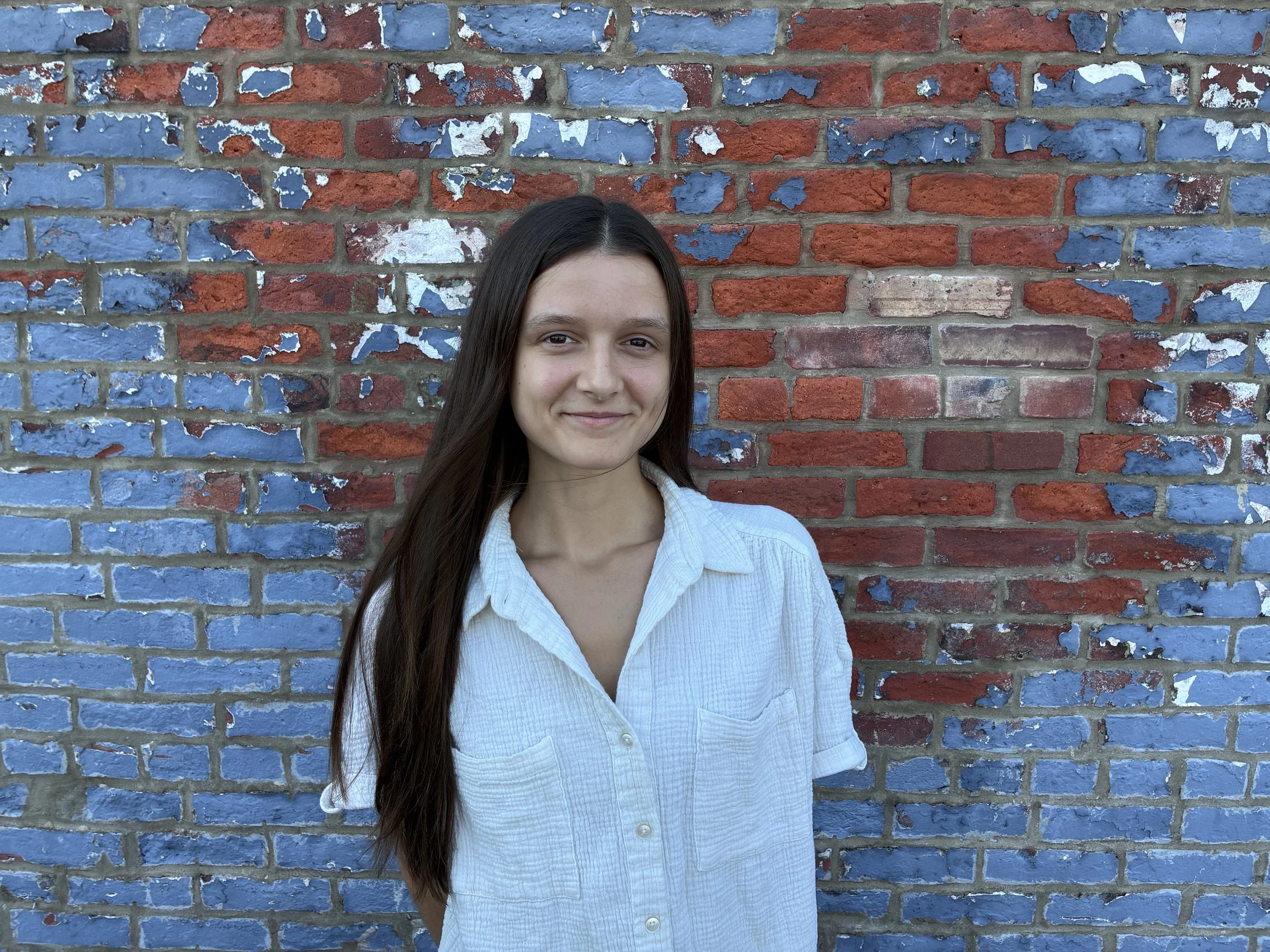 A young woman with long dark hair wearing a white button-up shirt standing in front of a brick wall painted with patches of blue and white paint.