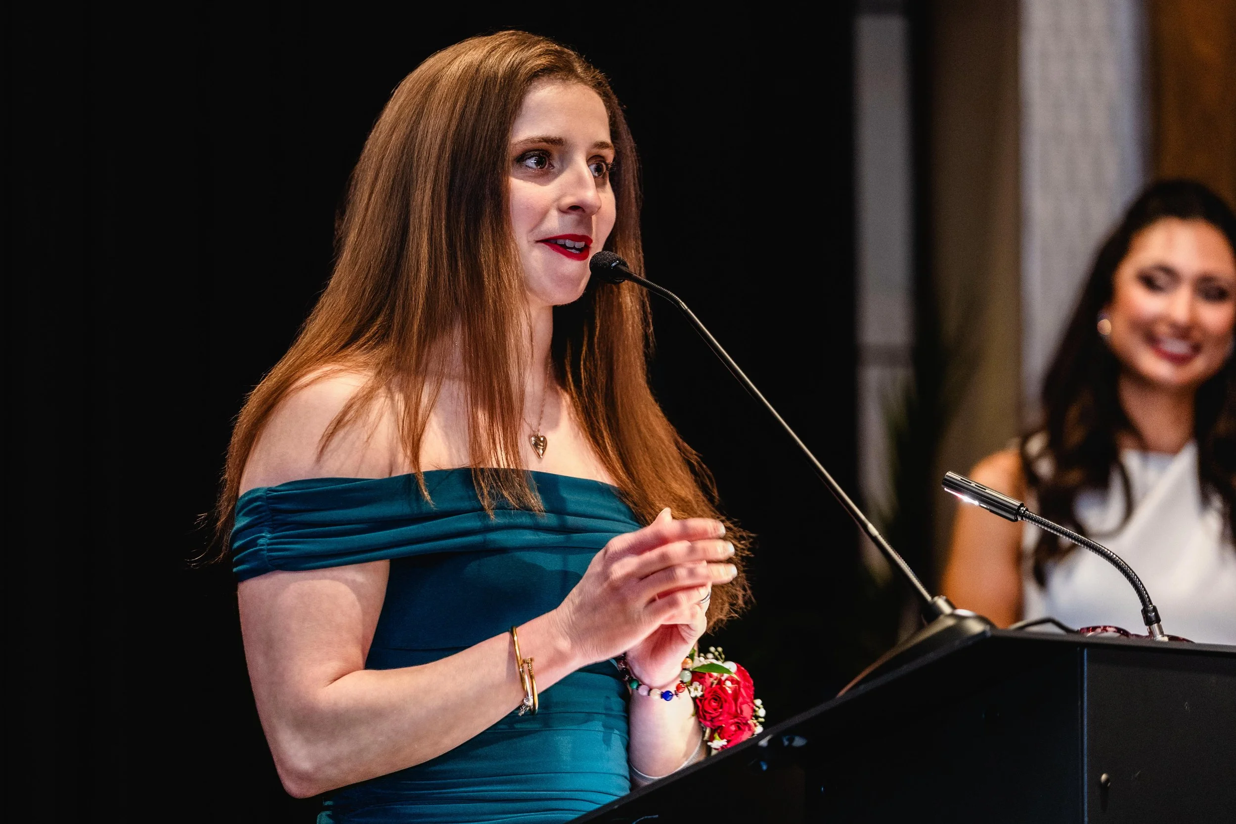 A woman with long red hair wearing a teal dress speaking at a podium with a microphone, with another woman smiling in the background.