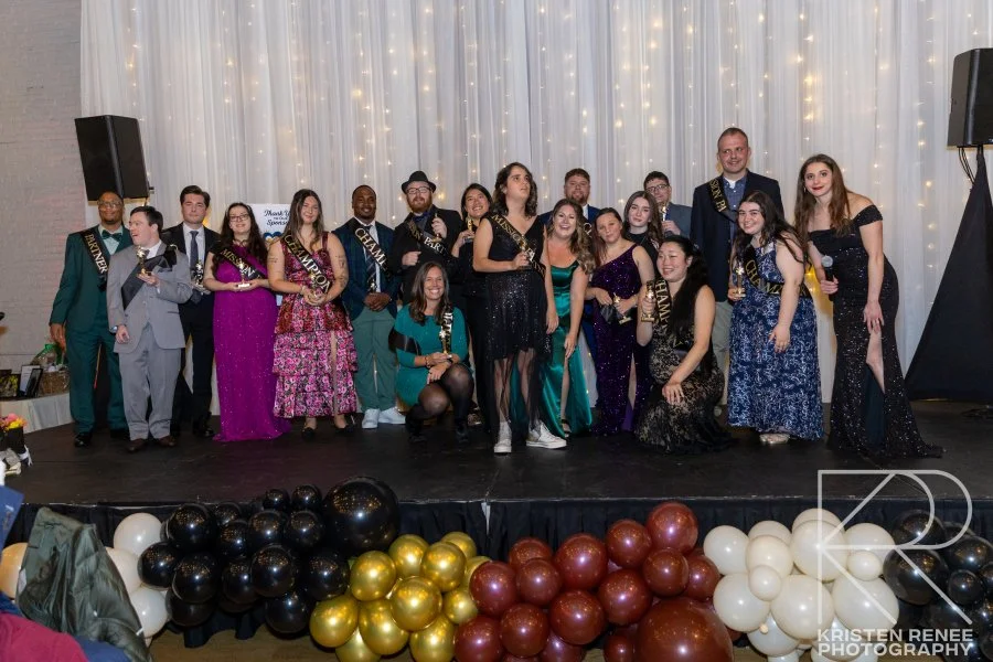 Group of young adults on stage at a prom event, wearing sashes and formal attire, with balloons in black, gold, red, and white at the front, and a curtain backdrop with string lights.
