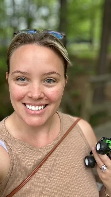 A smiling woman taking a selfie outdoors in a wooded area, holding a small black camera.