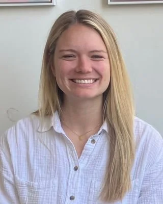 A smiling young woman with long blonde hair, wearing a white button-up shirt, standing indoors against a light-colored wall.