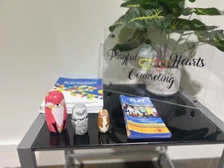 Decorative pink and white owl figurine, a gray and white skull figurine, a brown and white dog figurine, and a stack of brochures on a black table with a potted plant and a sign that reads "Playful Hearts Counseling" in the background.