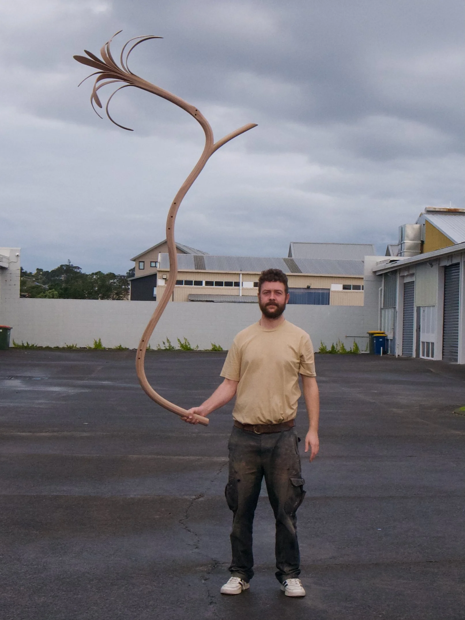 A man standing outdoors on a paved area, holding a large, curved wooden sculpture resembling a stylized flower or plant, with a cloudy sky overhead and buildings in the background.