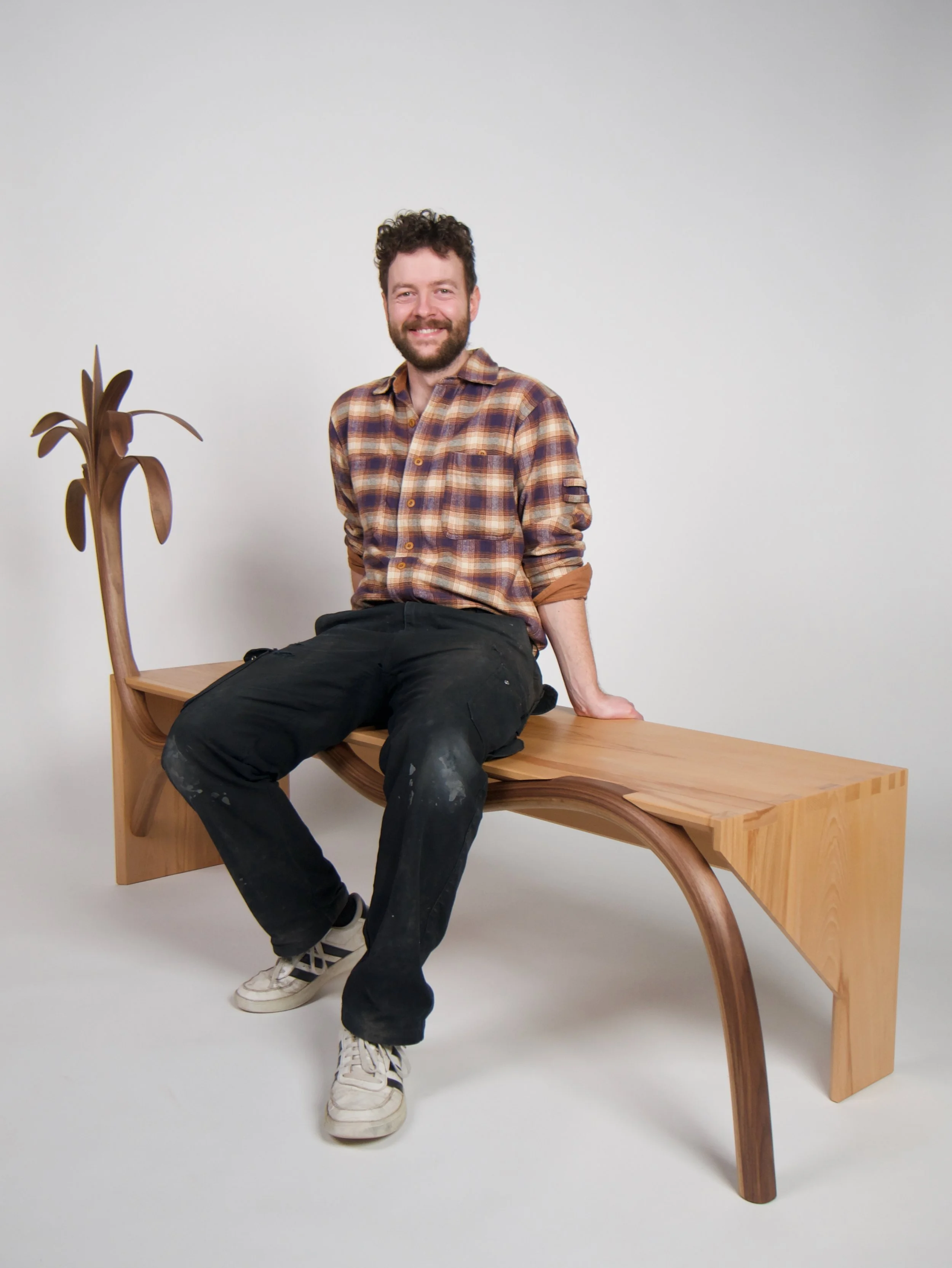 A man with a beard and curly hair sitting on a wooden bench with a flower-shaped backrest, smiling at the camera, against a plain white background.