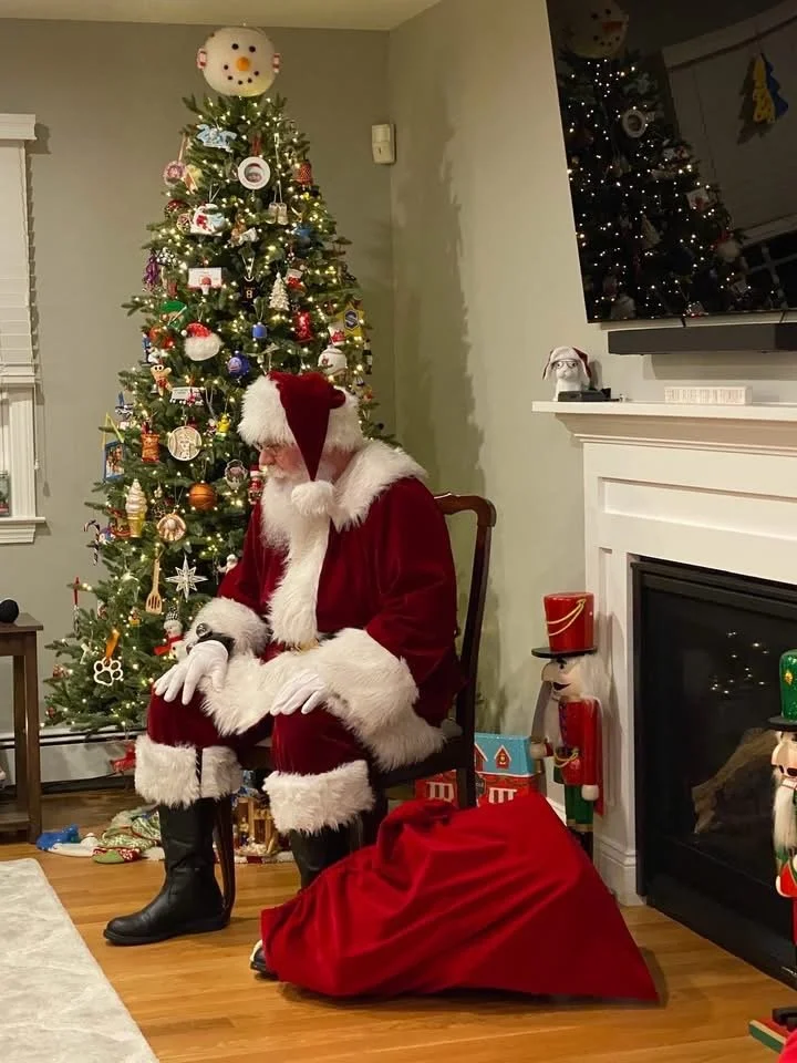 Santa Claus sitting in a chair next to a decorated Christmas tree, with gifts around. Santa is dressed in a traditional red suit with white trim, wearing a Santa hat, and black boots. A large red sack lies on the floor, likely filled with presents.