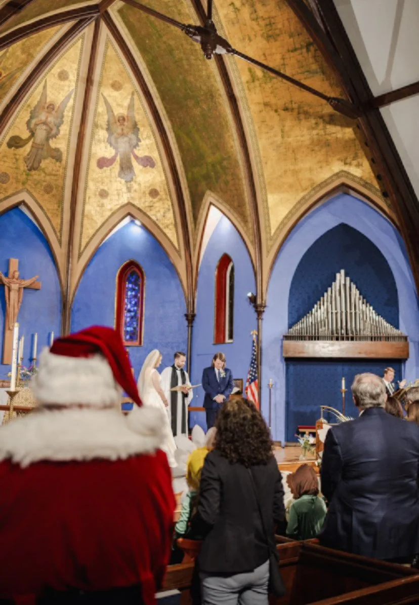 A church interior decorated for Christmas with a festive Santa hat in the foreground, people attending a wedding or ceremony, a large cross, stained glass windows, and a pipe organ on the wall.