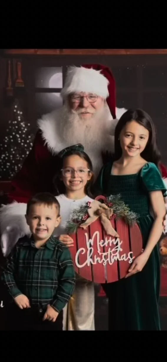 A family photo with Santa Claus, two young girls, and a young boy, holding a sign that says 'Merry Christmas' on a festive background with Christmas trees and lights.
