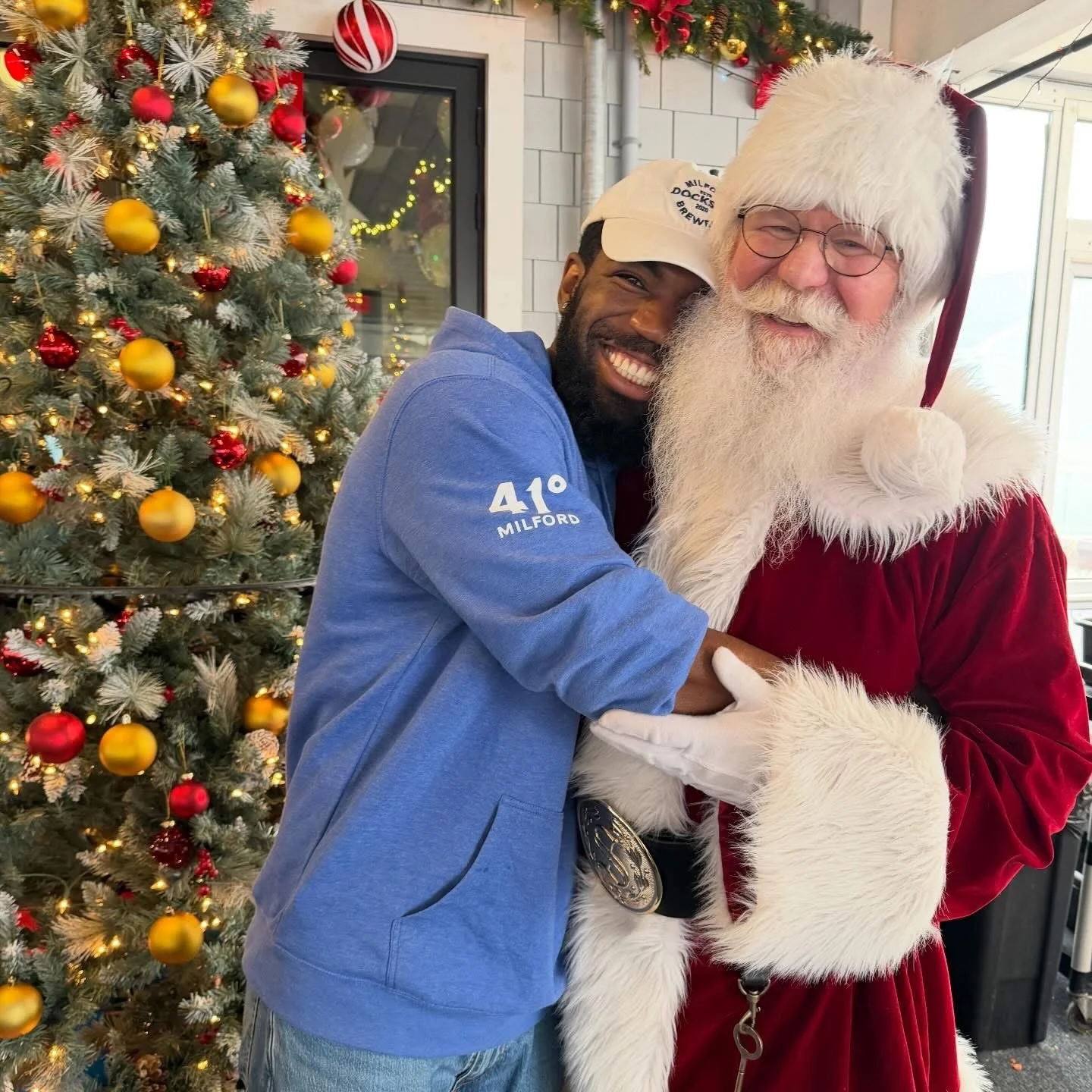 A man with a beard and a Santa Claus dressed person hugging and smiling in front of a decorated Christmas tree with yellow and red ornaments.
