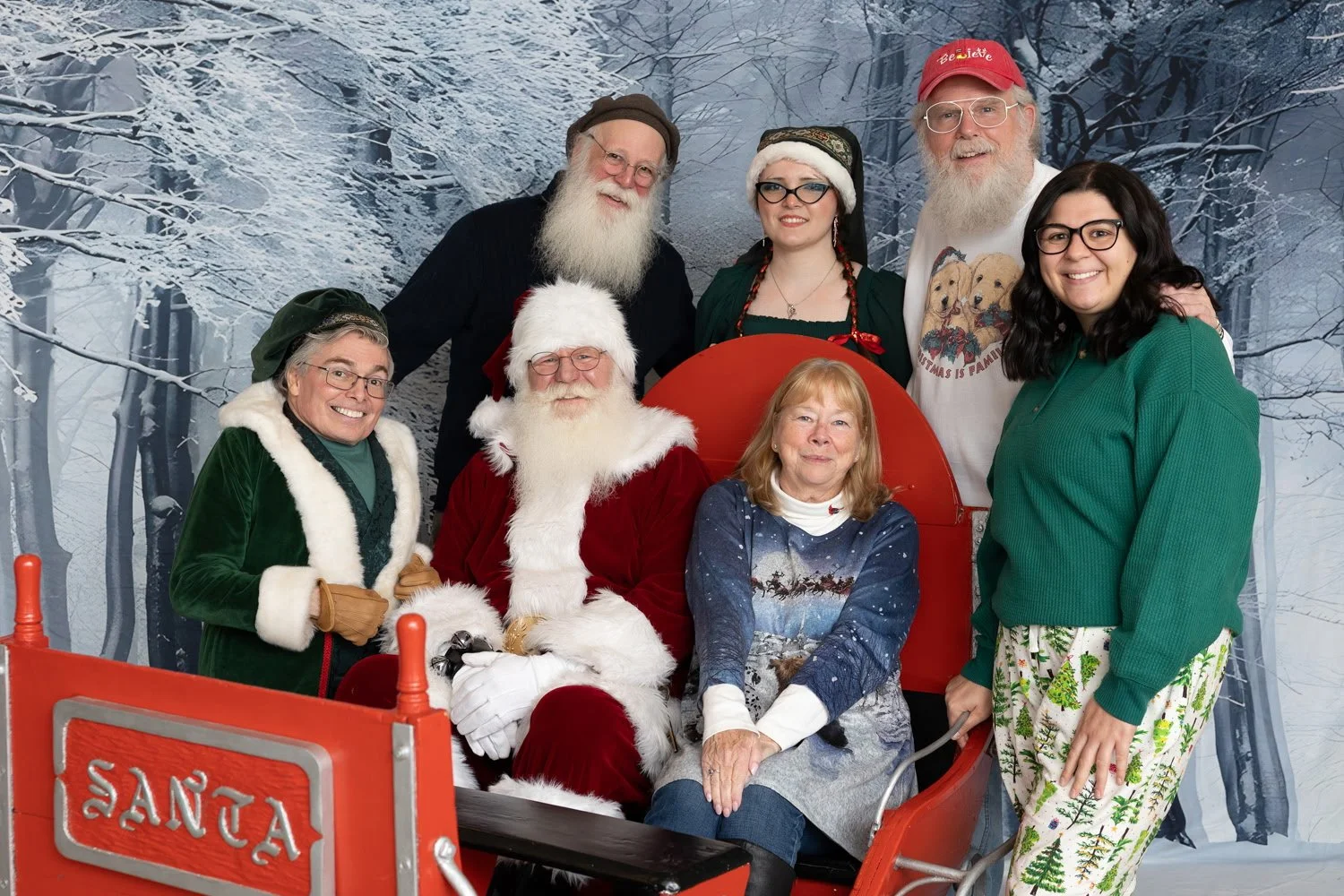 A group of eight people posing with Santa Claus in a snowy forest backdrop. Santa is sitting in a sleigh with a woman on his lap. The group includes individuals dressed in holiday-themed clothing, some wearing glasses and Santa hats.