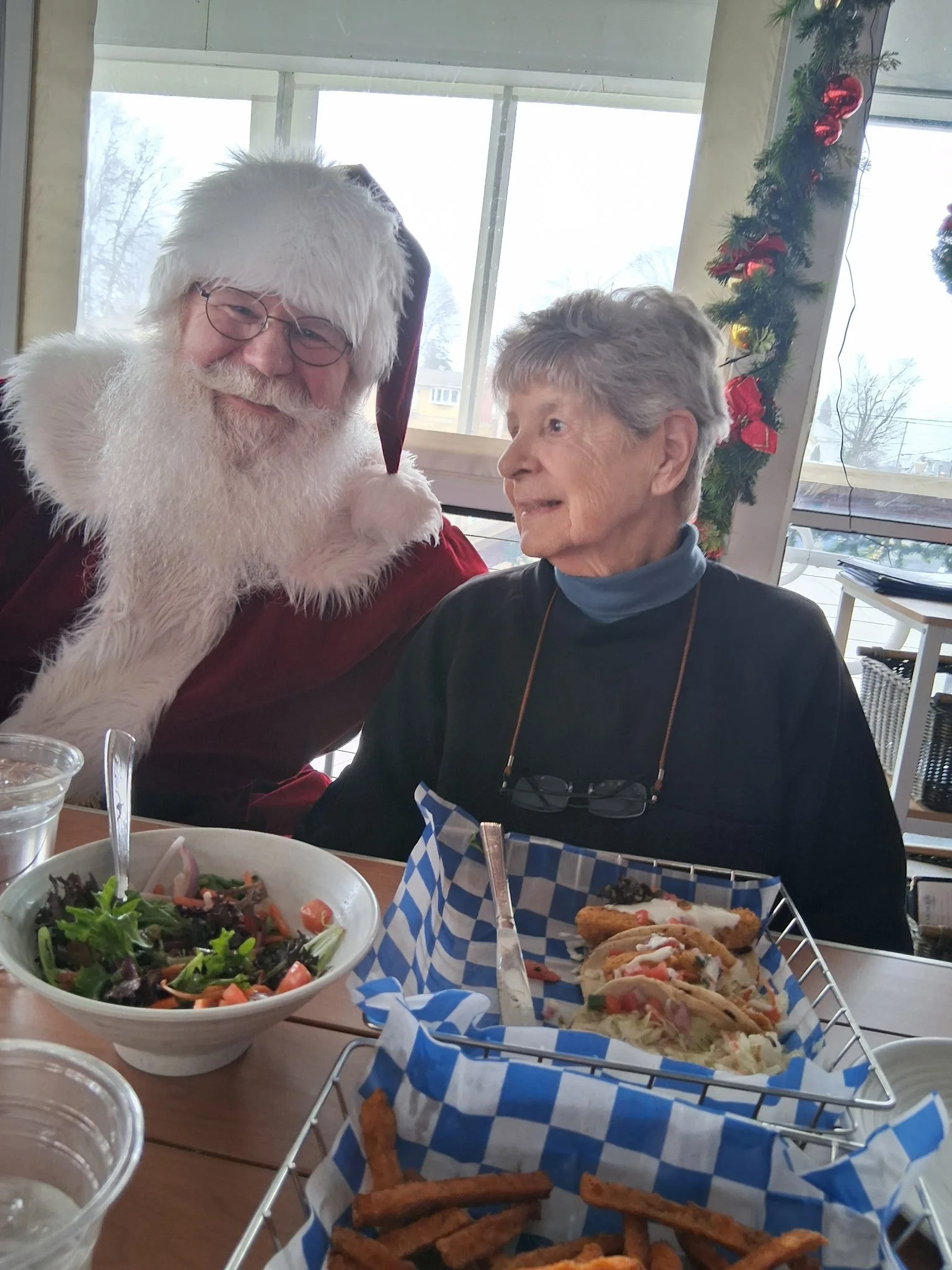 A man dressed as Santa Claus smiling and an elderly woman sitting at a dinner table decorated for Christmas with a Christmas garland on the window. The table has a salad, pizza, and fries.