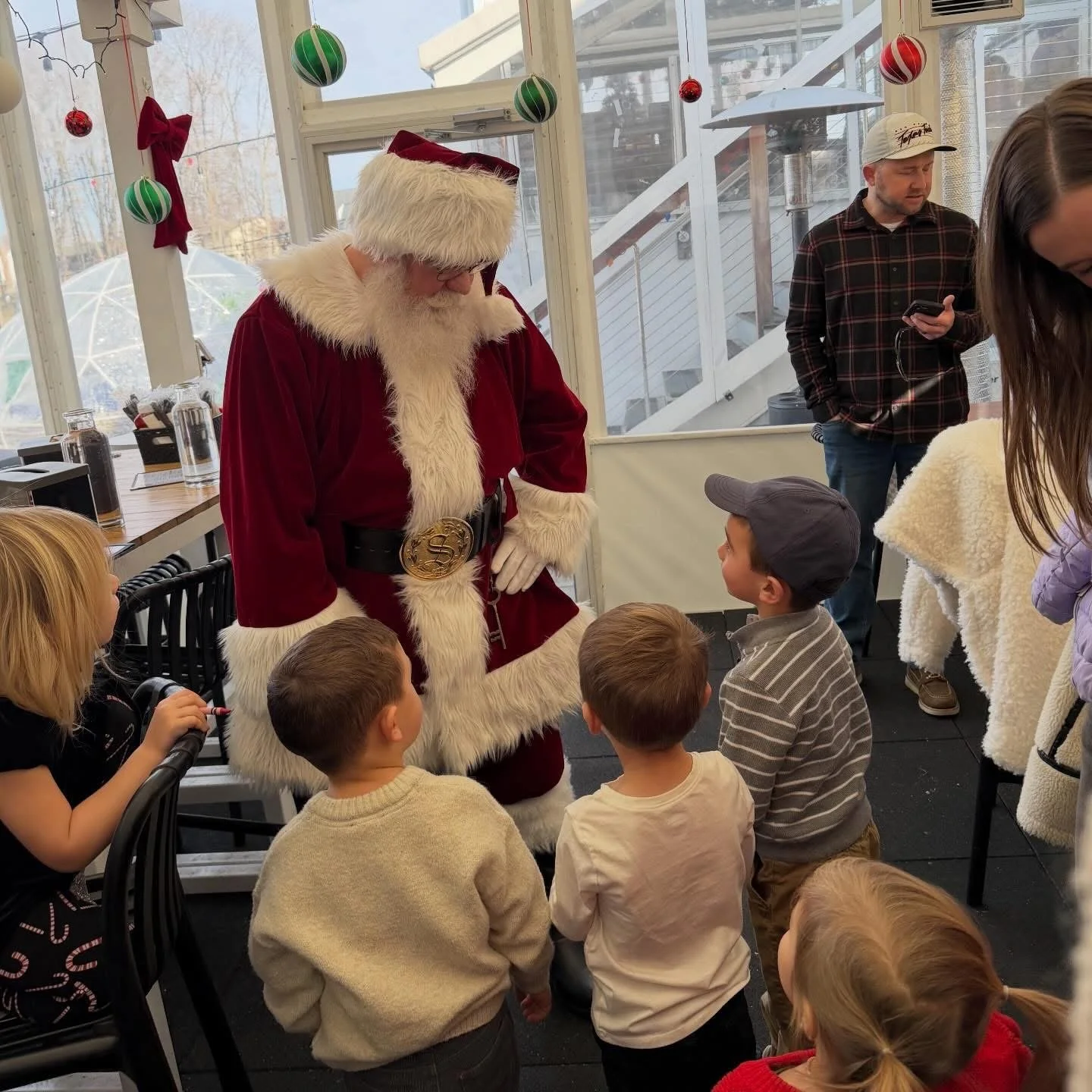 Santa Claus, dressed in a red velvet suit with white fur trim, is talking to a group of children inside a festive decorated room with Christmas ornaments hanging from the ceiling and windows. Two adults are standing nearby, one using a phone and the 