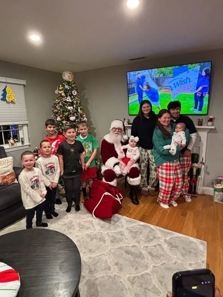 Family gathering with children and a person dressed as Santa Claus in a living room decorated for Christmas, with a Christmas tree, holiday decorations, and a television showing a scene outside.