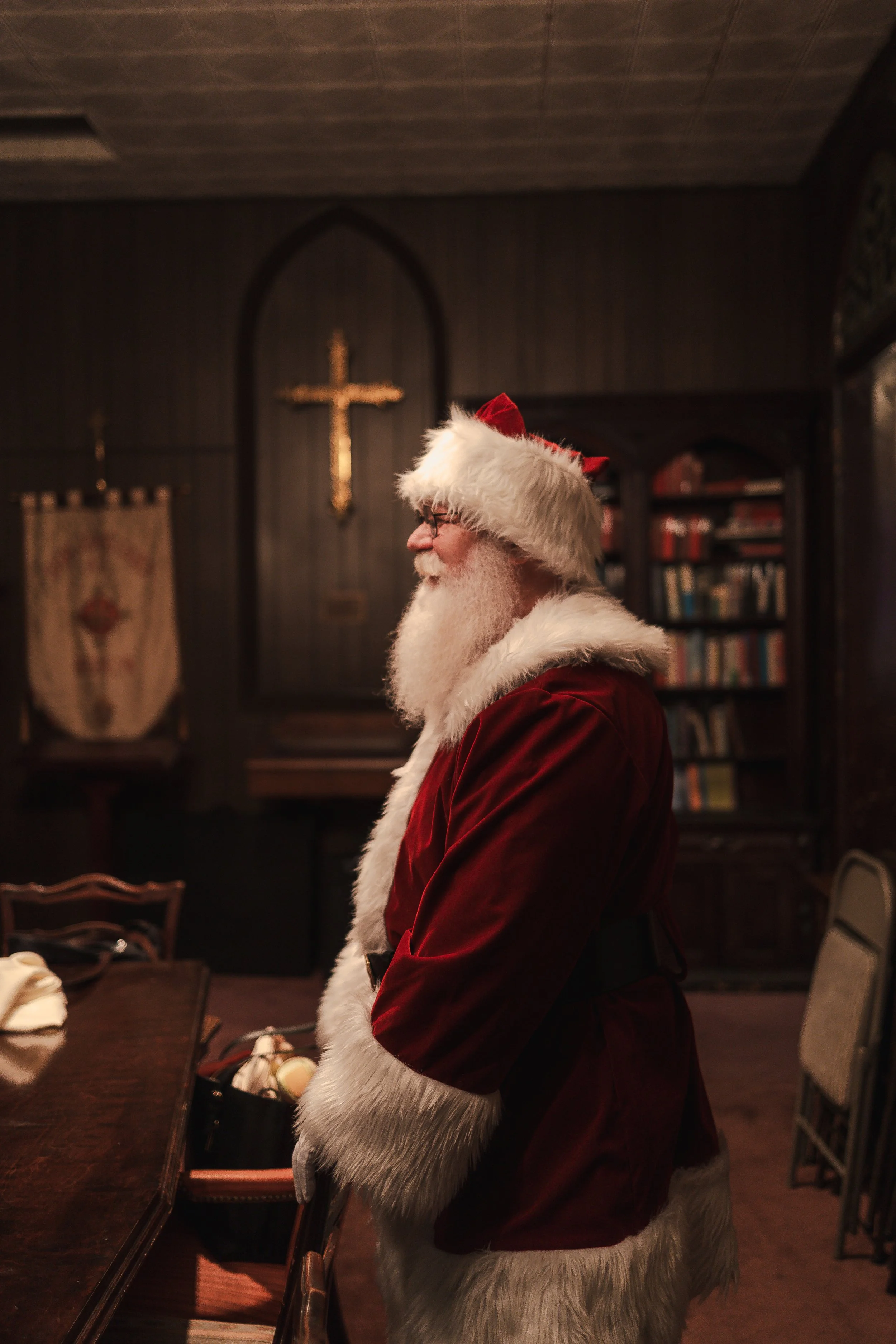 Santa Claus in a red velvet suit with white fur trim and a red hat with white fur, standing inside a dimly lit room with wooden walls and a cross hanging on the wall in the background.