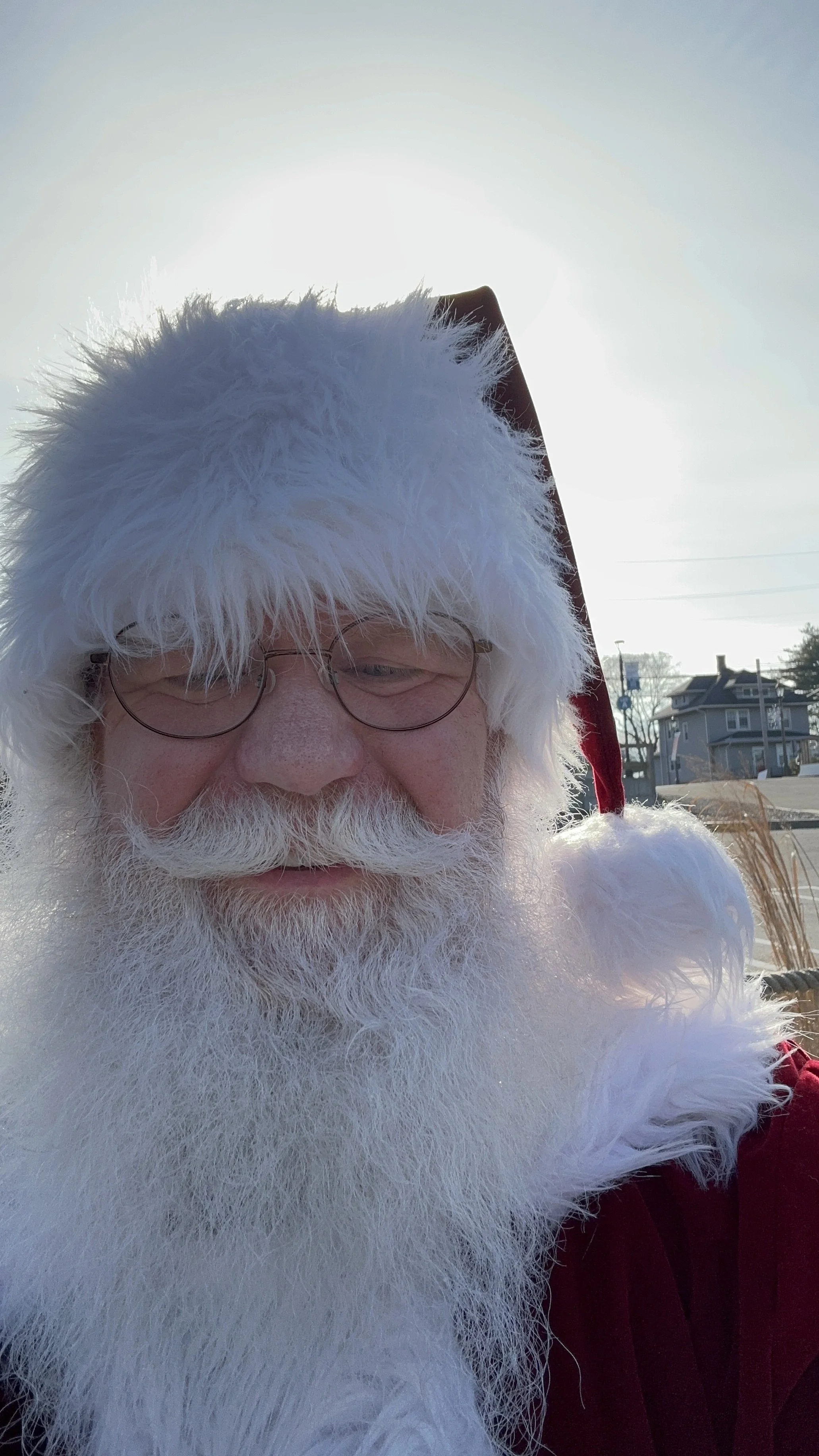 A close-up photo of Santa Claus outdoors with a bright sky behind him, wearing glasses and a traditional red Santa suit with white fur trim.