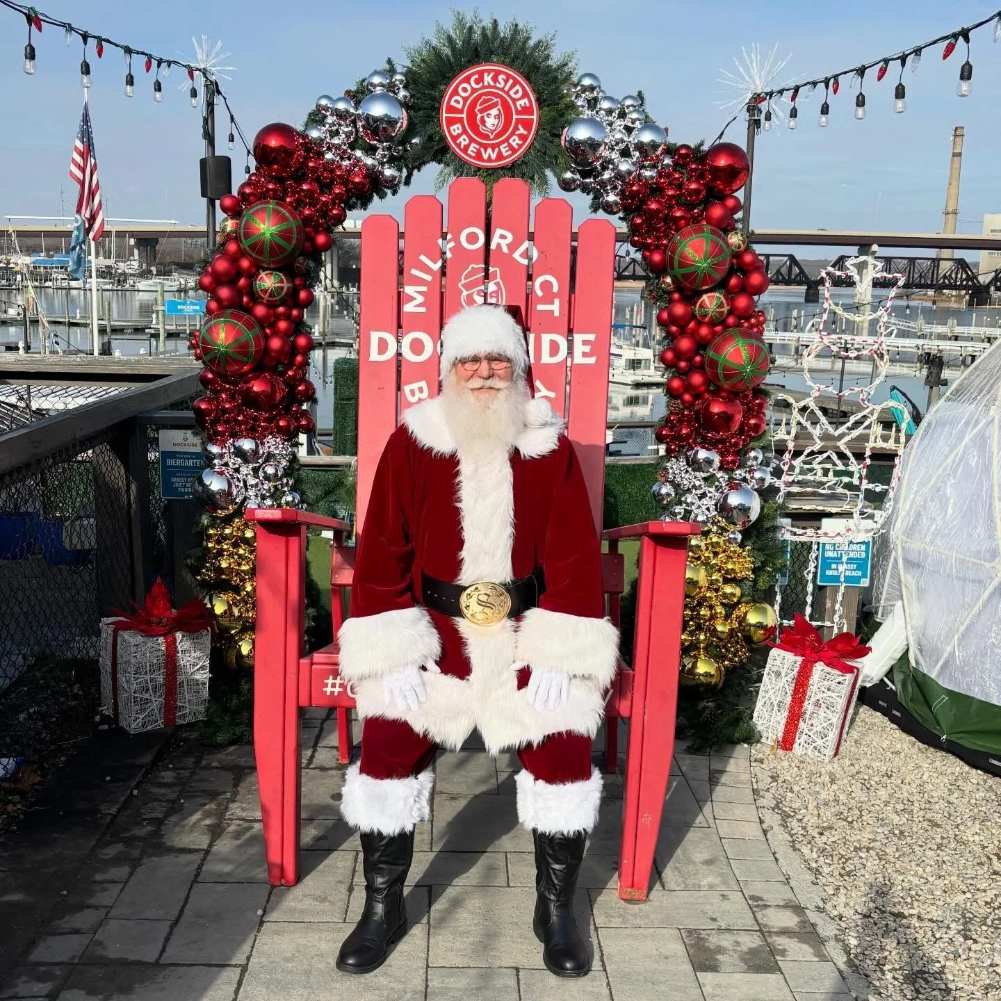 A person dressed as Santa Claus sitting on a large red chair surrounded by Christmas decorations, including a decorated arch with red and silver ornaments and a sign that reads 'Middle of the Dock'. In the background, there is a marina with boats and